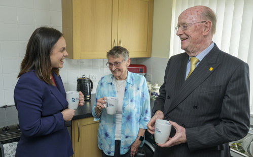 DFM Kate Forbes, Muriel Allison, Russel Griggs standing in Muriel's kitchen having a cup of tea and laughing